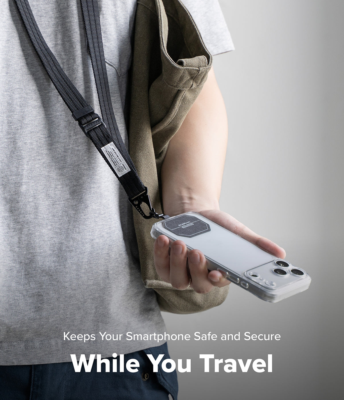 Holder Link Tactical Webbing Lanyard Strap - A person wearing a gray t-shirt and khaki jacket holds a smartphone in a clear case with a black magnetic strap attached, shown against a neutral gray background with text overlay 'Keeps Your Smartphone Safe and Secure While You Travel'.