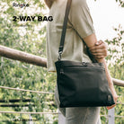 2-Way Bag - A person wearing a black crossbody bag with a zippered front pocket outdoors against a blurred green background.
