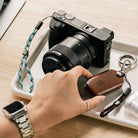 Paracord Lanyard Wrist Strap [6 Pack] - A hand holds a leather key fob near a camera with a blue and white paracord strap.